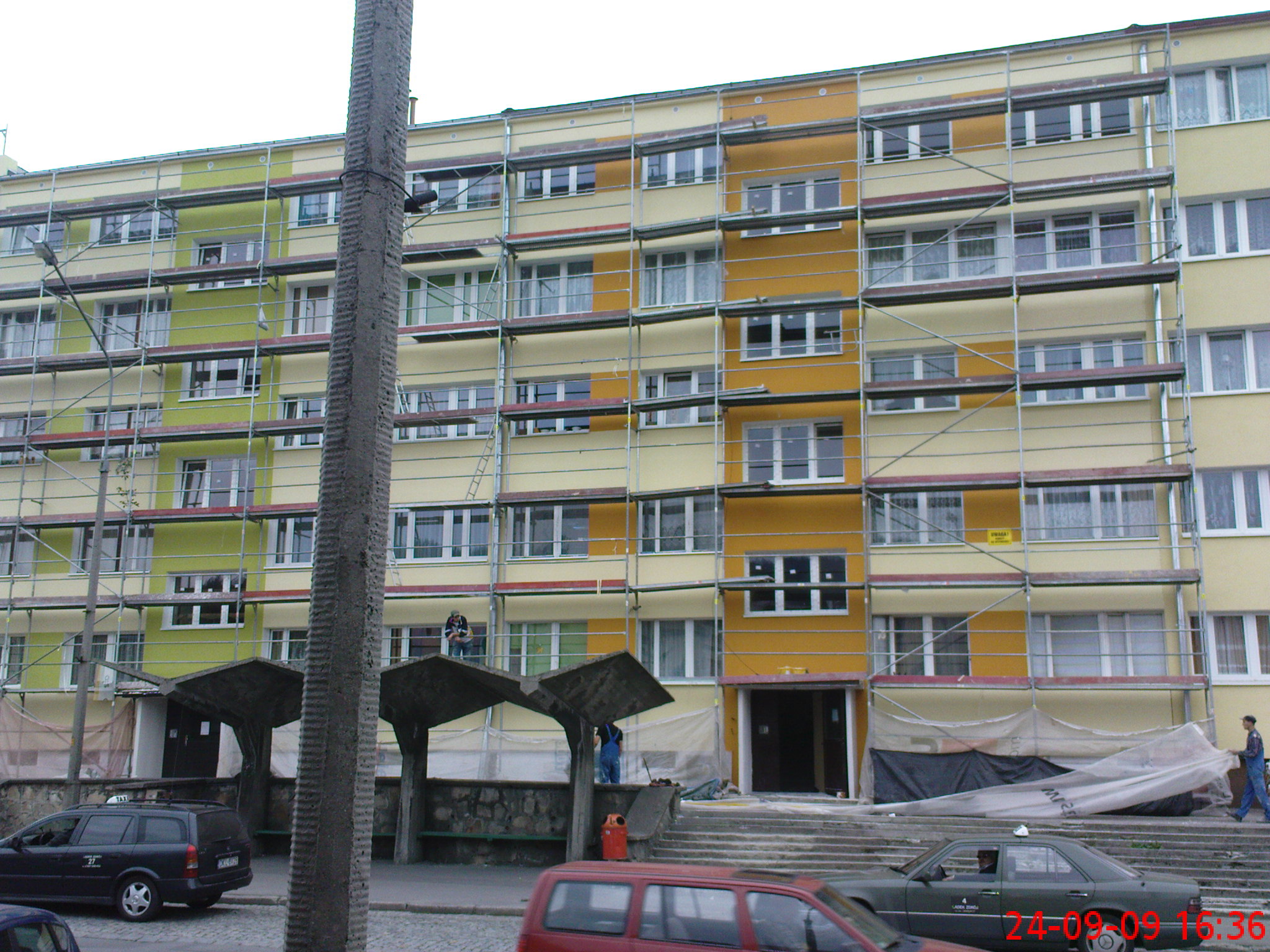 Wielopiętrowy budynek w trakcie docieplania elewacji, otoczony rusztowaniami, z widocznymi pracownikami na rusztowaniach i osłoniętym wejściem.